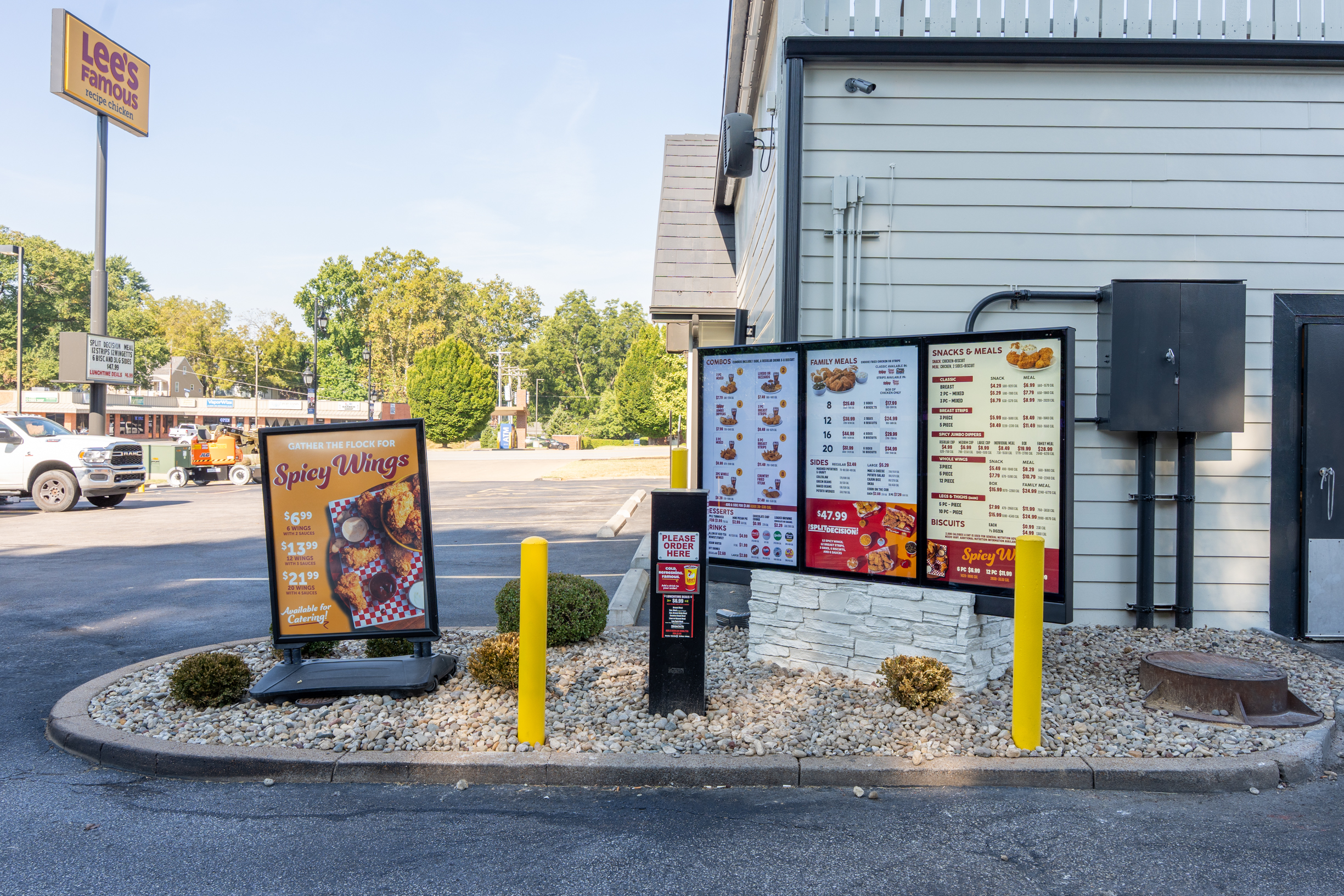 Exterior drive-thru area of a Lee's Famous Recipe Chicken restaurant in St. Charles. A freestanding promotional sign in the foreground advertises Spicy Wings, with pricing for 4, 8, and 16 wings and a note that they're available for catering. Behind it, a large illuminated digital menu board displays combo meals, family meals, and a snacks and meals section. A black ordering kiosk stands in the center. Yellow safety bollards line the drive-thru lane, and the Lee's Famous Recipe Chicken pole sign is visible in the background against a clear blue sky.