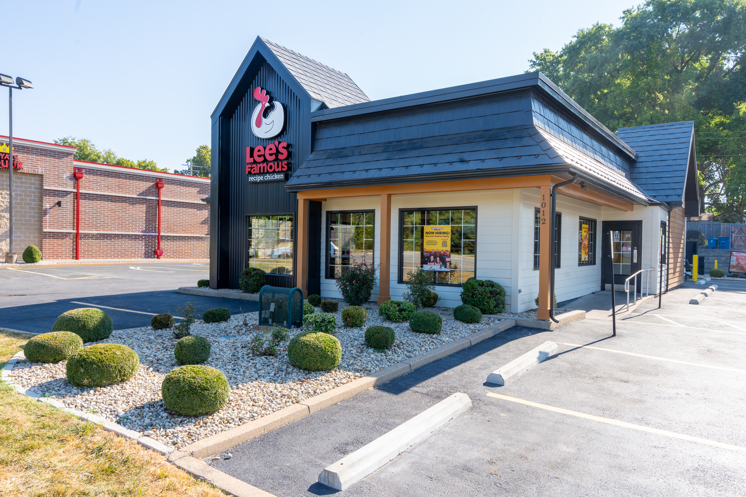 Exterior view of a Lee's Famous Recipe Chicken restaurant in St. Charles, featuring the brand's modern black-and-white barn-style facade with the red chicken logo, neatly landscaped shrubs, and a "Now Hiring" sign in the front window.