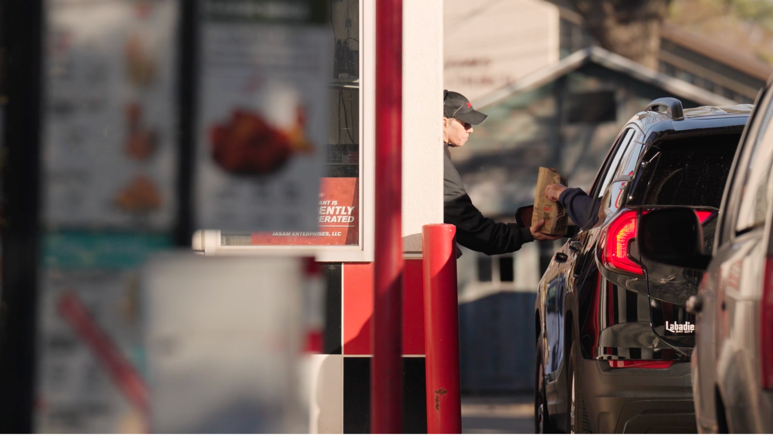 Fast food drive-thru worker handing a paper bag to a customer in a dark SUV, with red drive-thru pillars and a blurred menu board visible in the background.